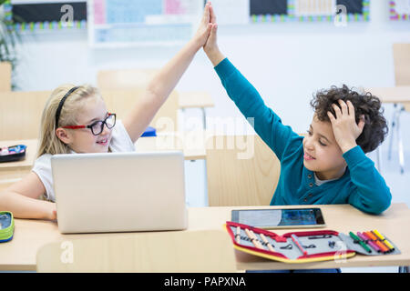 Schoolboy and schoolgirl with laptop high fiving in class Stock Photo