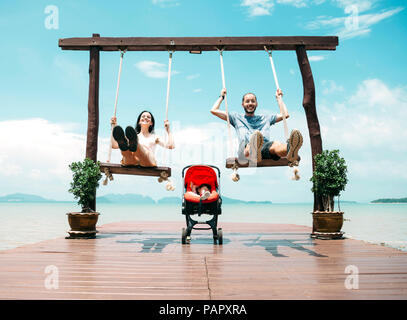 Thailand, Koh Lanta, happy parents  on wooden swings in front of the sea and sleeping baby in a stroller Stock Photo