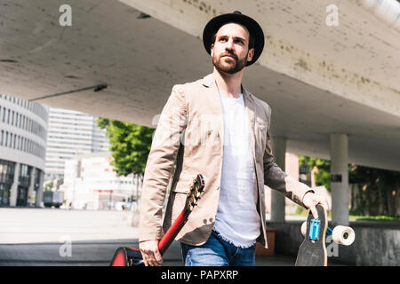 Young man with guitar and skateboard walking in the city Stock Photo