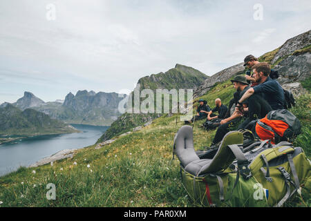 Norway, Lofoten, Moskenesoy, Group of young men sitting on grass, looking over Kjerkefjord Stock Photo