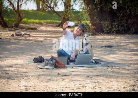 Smiling woman sitting on blanket on beach with dog taking a selfie Stock Photo