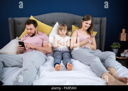 Neglected little girl sitting on bed with her parents, using smartphones Stock Photo