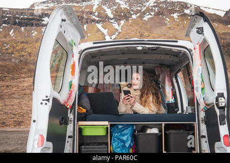 Portrait of young woman lying in van using smartphone and laptop Stock Photo
