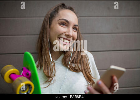 Portrait of smiling teenage girl with cell phone, earphones and skateboard Stock Photo