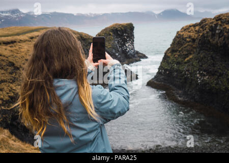 Iceland, back view of young woman taking picture with smartphone at coast Stock Photo