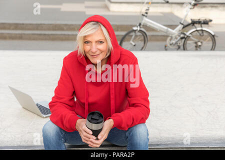 Senior woman wearing red hoodie sitting outdoors with laptop and takeaway coffee Stock Photo