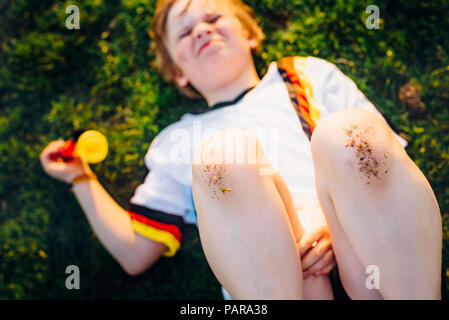 Boy in German soccer shirt lying on grass, with dirty knees Stock Photo