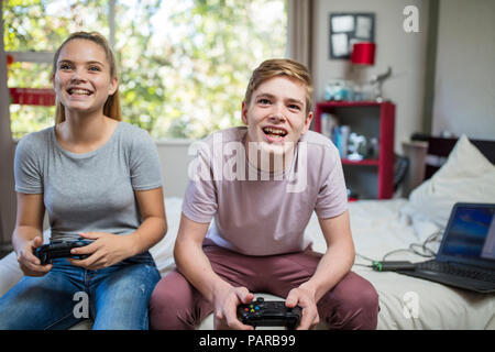 Happy teenage girl and boy sitting on bed playing video game