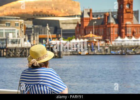Woman wearing sun hat leaning on railings looking out over Cardiff Bay towards landmark buildings on the waterfront. Stock Photo