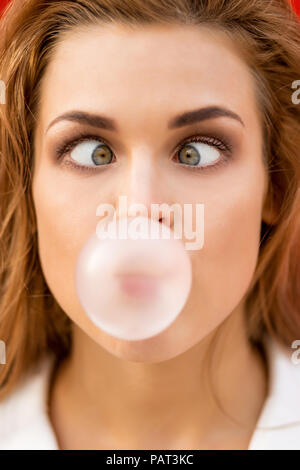 Funny portrait of beautiful young cute woman in white vogue suit look to her nose while blowing gum bubble against red wall. Urban style girl, quick l Stock Photo