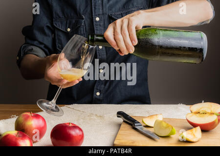 Man pours a glass of vintage apple wine out of ice cold bottle. Male hands pouring premium cidre in wine glass above rustic wood table. Stock Photo