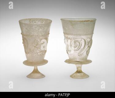 Goblets with Applied Decoration. Date: 11th-early 12th century.  These matcing goblets are each formed by a conical cup attached to a small, solid, and splayed stemmed foot by a circular flange applied around the base of the cup. They are made of yellowish colorless glass that contains many small bubbles. Both cups are decorated with an unbroken applied trail in the same yellowish color, which forms a horizontal line about two-thirds of the height and continues below to create a fanciful, abstract pattern of curly designs around the cup. The decoration can be read more clearly when the cup is 