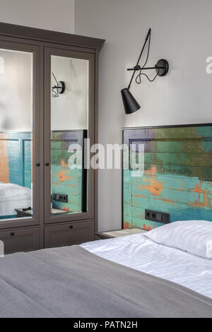 Nice room in a hotel with light walls. There is a bed with a colorful wooden bedhead, white linens with a coverlet on the bed, nightstand, black lamp  Stock Photo