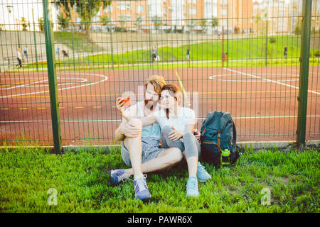 A mans pair red hair and beard and woman rest after playing sports outdoors on grass in background of stadium. The guy holds red smartphone on outstre Stock Photo