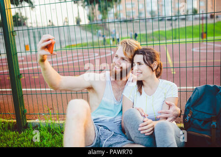 A mans pair red hair and beard and woman rest after playing sports outdoors on grass in background of stadium. The guy holds red smartphone on outstre Stock Photo