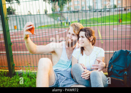 A mans pair red hair and beard and woman rest after playing sports outdoors on grass in background of stadium. The guy holds red smartphone on outstre Stock Photo