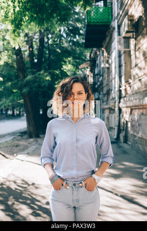 Happy young woman with naturally curly hair standing in city looks at camera. Vertical candid image of fashionable female keeps hands in pockets and s Stock Photo