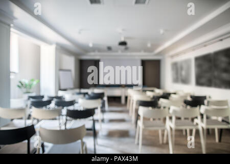 Blurred photo of a conference hall with white walls and a gray floor. There are many light and dark chairs, board, projector, tables, lamps, plant in  Stock Photo