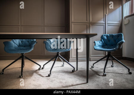 Empty meeting room with a gray carpet on the floor. There is a wooden table with three blue soft chairs, lockers and a window with a curtain. Horizont Stock Photo