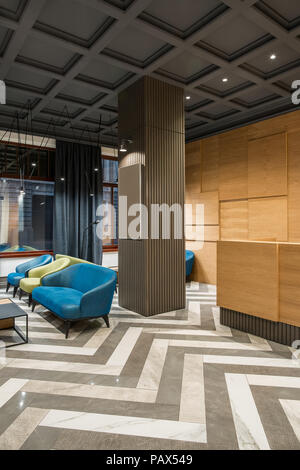 Fine interior with luminous lamps in the hotel with dark ceiling and patterned tiled floor. There is a wooden reception, column, multicolored armchair Stock Photo