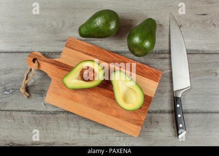 Table top view - avocado cut in half on wooden chopping board, chef knife next to it, two whole green pears above. Stock Photo