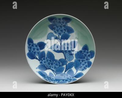 Dish with Hydrangeas. Culture: Japan. Dimensions: H. 2 1/4 in. (5.7 cm); Diam. 8 in. (20.3 cm). Date: ca. 1690-1730.  This dish draws the viewer's attention to a single sprig of hydrangea, a typical summer blossom of Japan, framed by a pale green mist. The flowers painted in cobalt blue paired with the hazy celadon glaze give the impression that we are seeing these flowers shortly after a summer rain shower.  The refined, almost poetic nature of this dish is appropriate considering its use. This is an example of Nabeshima ware, a specialty porcelain that was produced during the Edo period. The Stock Photo