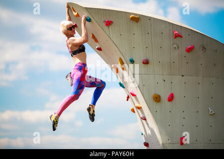 Photo from side of sports blonde in leggings hanging on wall for climbing against blue sky with clouds in afternoon Stock Photo