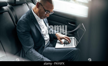 Businessman working on laptop computer while commuting to office in his sedan in back seat. Entrepreneur managing business work on the move sitting in Stock Photo