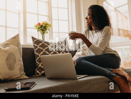 Businesswoman working on laptop computer sitting at home holding a coffee cup in hand. Smiling woman sitting on sofa at home and looking out of the wi Stock Photo