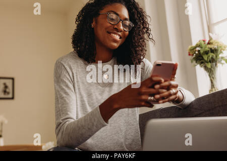 Smiling businesswoman sitting on sofa at home using mobile phone while working on laptop. Woman working on laptop computer at home and managing her bu Stock Photo