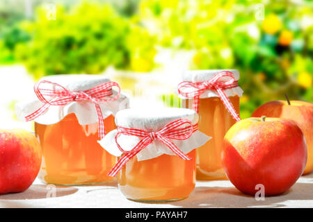 Homemade apple jelly in glass jars with linen cover and a nostalgic ribbon bow in bright sunshine with apple trees in the background Stock Photo