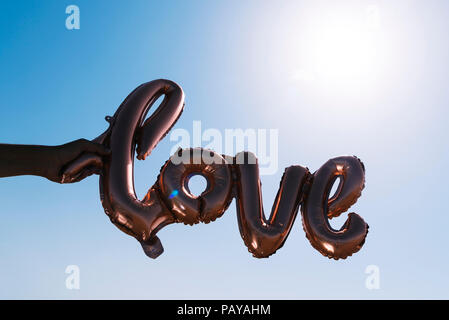 closeup of the hands of a young caucasian man holding a balloon in the shape of the word love against the sky, in backlit with a sunbeam in the backgr Stock Photo