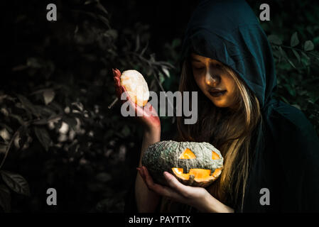 Witch opening pumpkin lid by hand. Old woman holding bright pumpkin in dark forest. Halloween day and Mystery concept. Fantasy of magic theme. Demon a Stock Photo