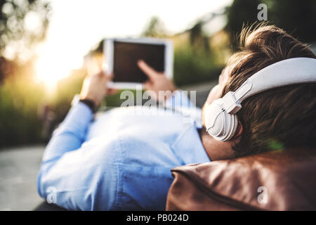 A businessman with tablet and headphones, lying on the bench outdoors at sunset. Stock Photo