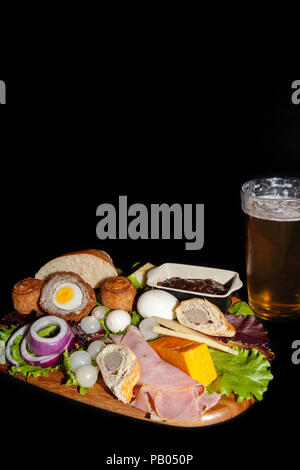 Traditional English pub meal of a ploughman’s lunch served with a pint of lager. The typical cold food includes pork pies, ham, hard boiled eggs and c Stock Photo
