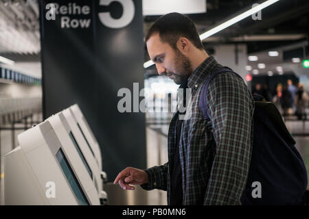 Young man with backpack touching interactive display using self service machine, doing self-check-in for flight or buying airplane tickets at automatic device in modern airport terminal building Stock Photo