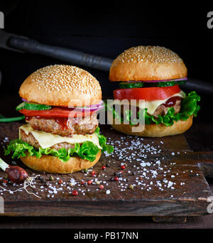 two burgers with a meat chop, vegetables and green lettuce in a round bun with sesame seeds on a brown wooden board, spices are scattered nearby Stock Photo