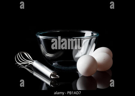 Glass transparent bowl with metal whisker and three white eggs aside from side on black background Stock Photo