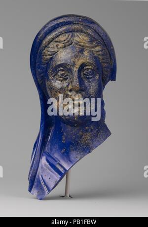 Glass portrait head of a woman. Culture: Roman. Dimensions: H.: 7 1/2 in. (19.1 cm). Date: 1st half of 2nd century A.D..  Semi-opaque streaky blue.  Hollow block pressed into an open mold with irregular tooling marks on inside and ground flat edges around top of head.  Shaped by mold and then details added by carving: long flowing headdress in two folds above hair, falling onto shoulder; hair parted at the center and arranged in flowing locks to either side; arched brows; well-defined eyes with eyelids and upward-looking pupils; straight nose with flaring nostrils; small mouth with pursed lips Stock Photo