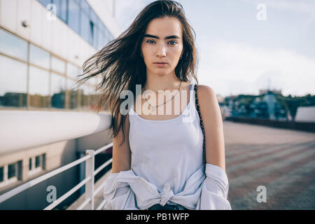 Beautiful young woman wearing white top posing at city street at summer day. Girl with fluttering long brown hair standing outdoors looking at camera. Stock Photo