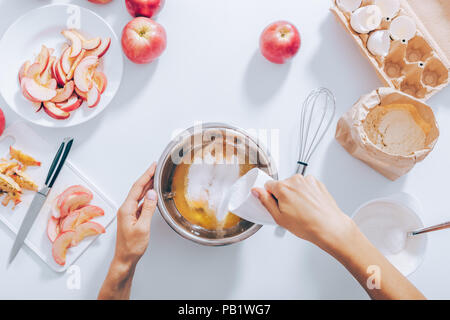 Process of making apple pie, top view. White table with ingredients for dough, cut and whole fruit. In the center female hand adding sugar to raw eggs Stock Photo