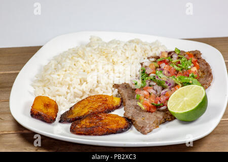 A steak covered in pico de gallo surrounded by plantains and white rice on a white plate. Cuban food. Stock Photo