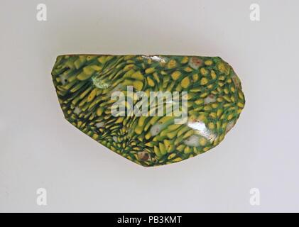 Glass mosaic bowl fragment. Culture: Roman. Dimensions: 1 1/2 × 2 1/16 × 1/8 in. (3.7 × 5.2 × 0.4 cm)  Estimated diameter: 3 9/16 in. (9.0 cm). Date: late 1st century B.C.-early 1st century A.D..  Translucent blue green appearing green, opaque yellow and white.  Vertical rim, slightly beveled inward; vertical side, then curving in sharply towards bottom  Mosaic pattern formed from polygonal sections of a single cane in a blue green ground with multiple yellow rods and a large central white rod.  Polished exterior; pitting of surface bubbles on exterior, including one large, deep hole below rim Stock Photo