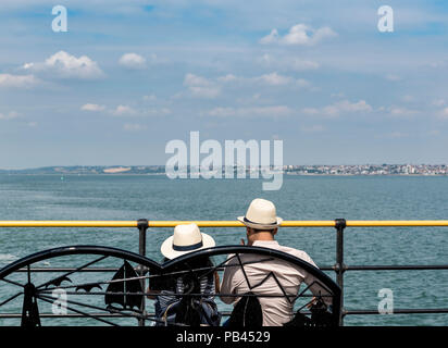 Young Asian couple sitting on a bench ovelooking the sea wearing straw hats. Stock Photo