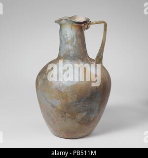 Glass jug. Culture: Roman. Dimensions: Overall: 5 3/4in. (14.6cm)  Other: 3 11/16in. (9.4cm). Date: mid-1st-2nd century A.D..  Translucent blue green; handle and trail in same color; purple streak in rim and neck.  Everted rounded rim, tooled into trefoil shape; funnel-shaped, oval neck; sloping shoulder; body tapering downward with convex sides; concave bottom; strap handle with three ribs applied to shoulder, drawn up and out, turned in horizontally to rim, and trailed on to top of neck and underside of rim.  Complete, but  cracks in upper body below handle, running up shoulder and neck to r Stock Photo