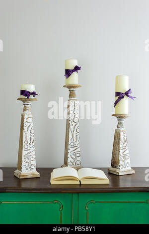 Three wooden candlesticks with engraved inscriptions holding three candles and opened old book with blank pages on green vintage wooden sideboard Stock Photo