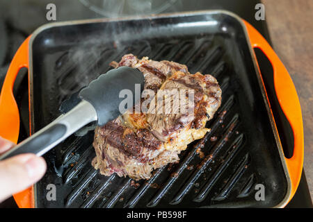 https://l450v.alamy.com/450v/pb598f/cooking-meat-on-cast-iron-grill-pan-indoors-at-home-person-turning-ribeye-steak-on-other-side-with-tongs-pb598f.jpg