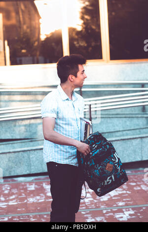 Student putting a notebook to a backpack. Boy standing at front of university building. Young boy wearing blue shirt and dark jeans Stock Photo