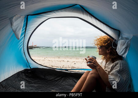 lonely beautiful woman sitting on the tent looking outside. wind on the hair and camping on the beach near the colors of water and shore. freedom and 