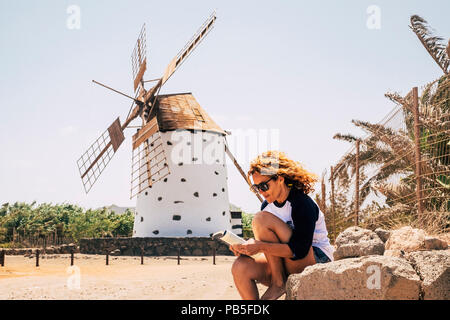 nice beautiful cheerful lady with blonde curly hair sitting under a windmill in outdoor scenic place reading a book and enjoying the leisure activity. Stock Photo